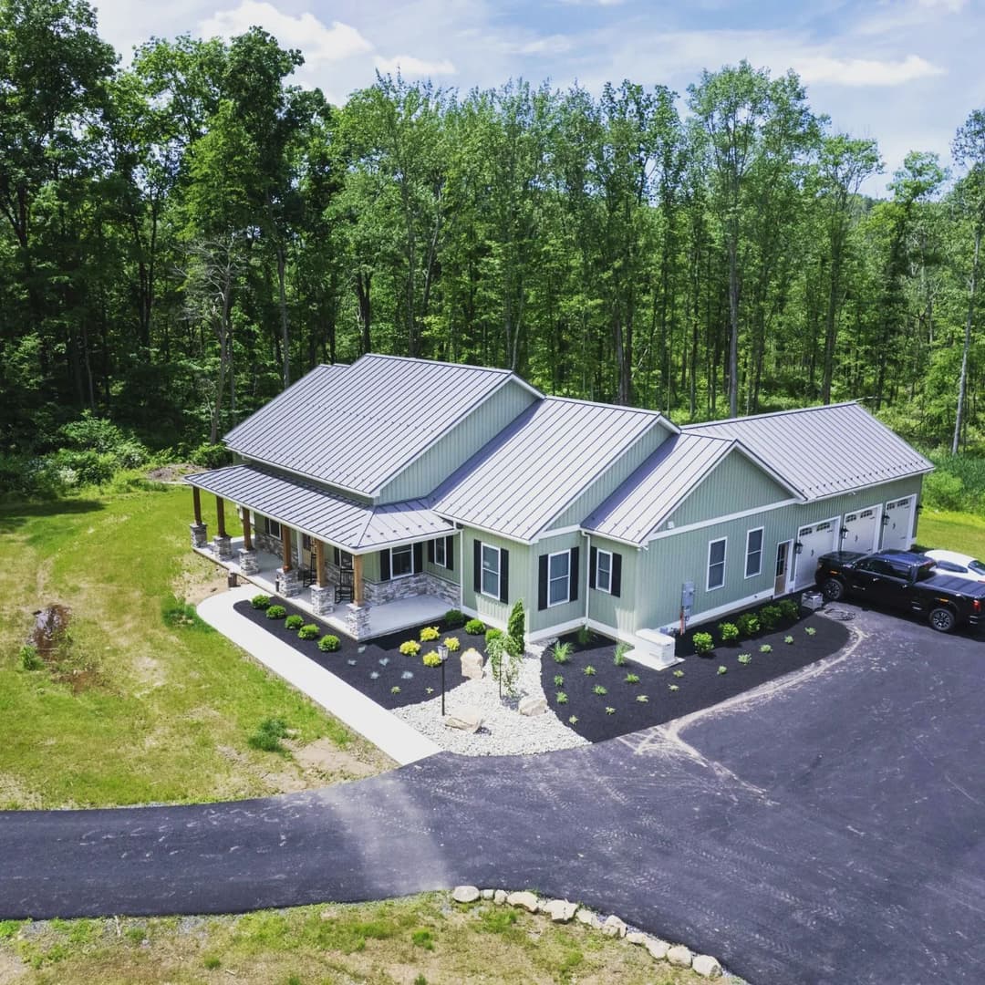 Aerial view of a modern green home with a gray metal roof, surrounded by trees and landscaping.