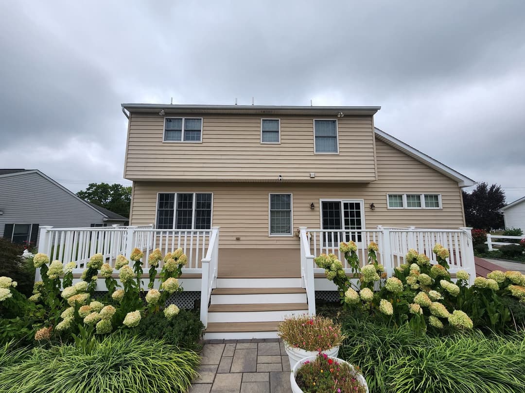 Two-story beige house with white railings, surrounded by green shrubs and flowers under a cloudy sky.