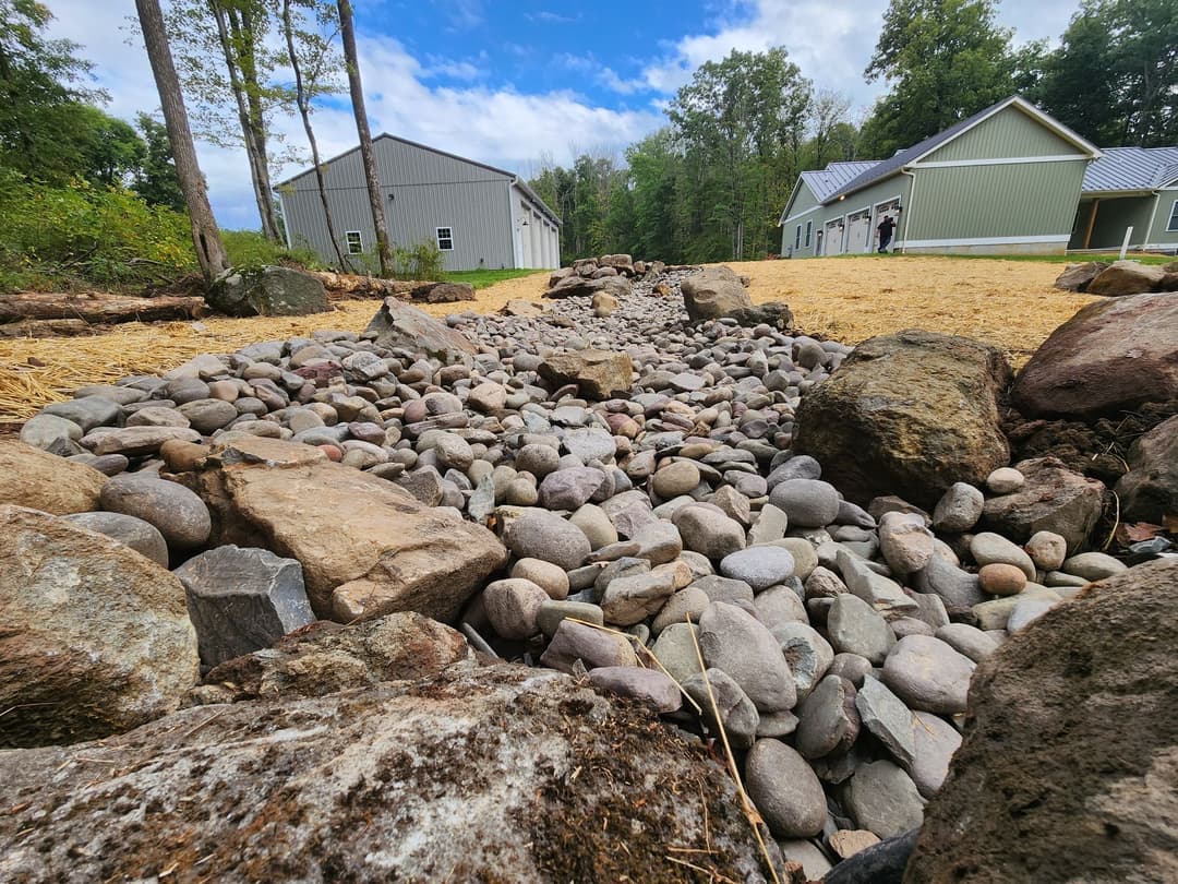 Stone pathway with gravel and rocks, surrounded by trees and buildings under a blue sky.