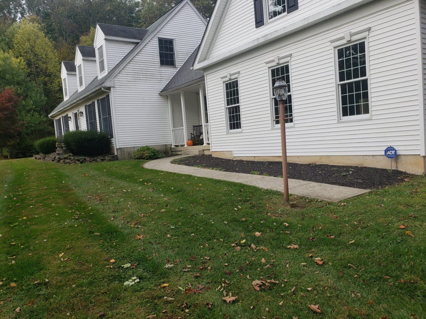 White two-story house with a manicured lawn and pathway, surrounded by trees and shrubs.