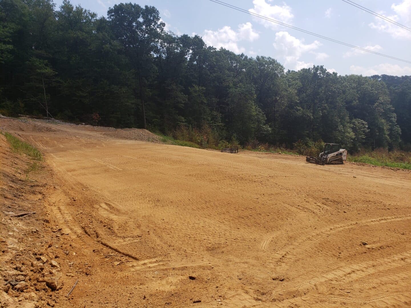 Cleared land with heavy machinery in forested area under blue sky and scattered clouds.