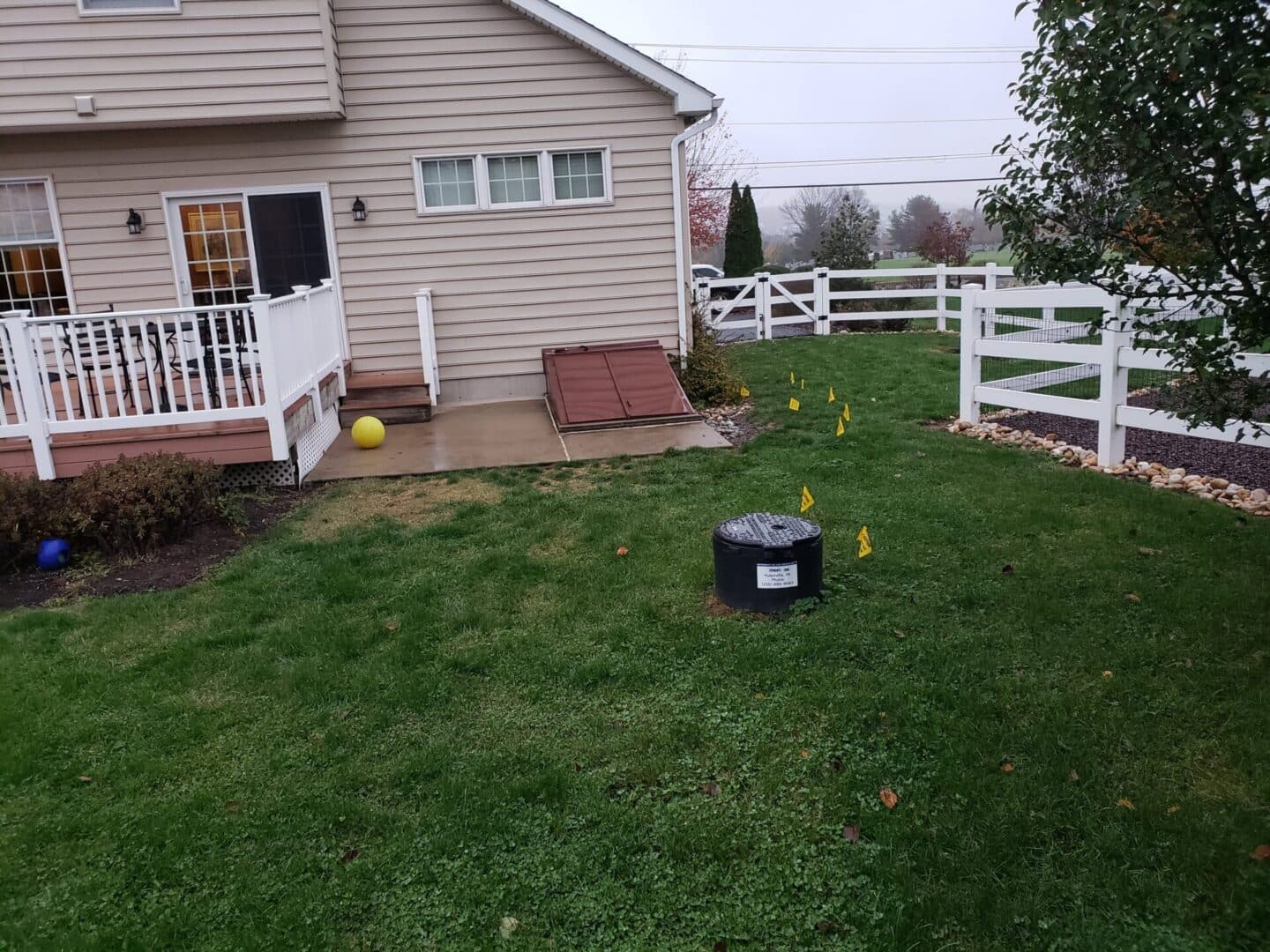 Backyard scene with a house, grassy area, yellow flags, and a drainage cover on a rainy day.