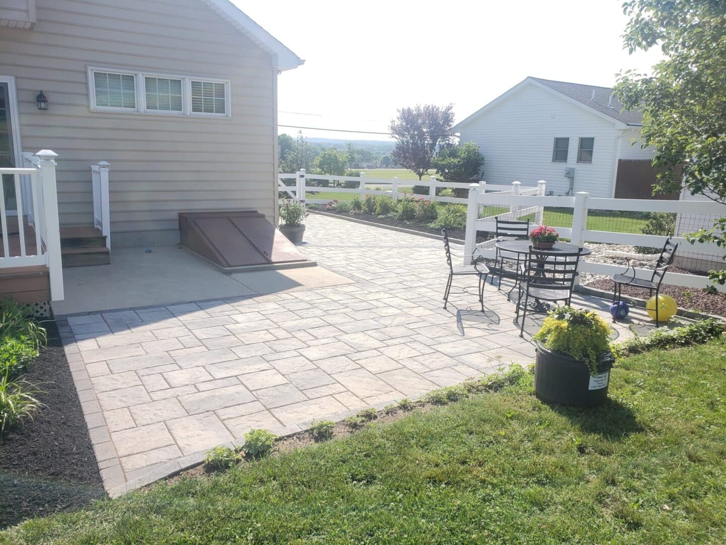 Paved patio with seating area and landscaped garden next to a house on a sunny day.