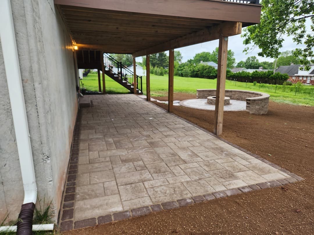 Patio with paver stones under a wooden deck, leading to a stone fire pit area.
