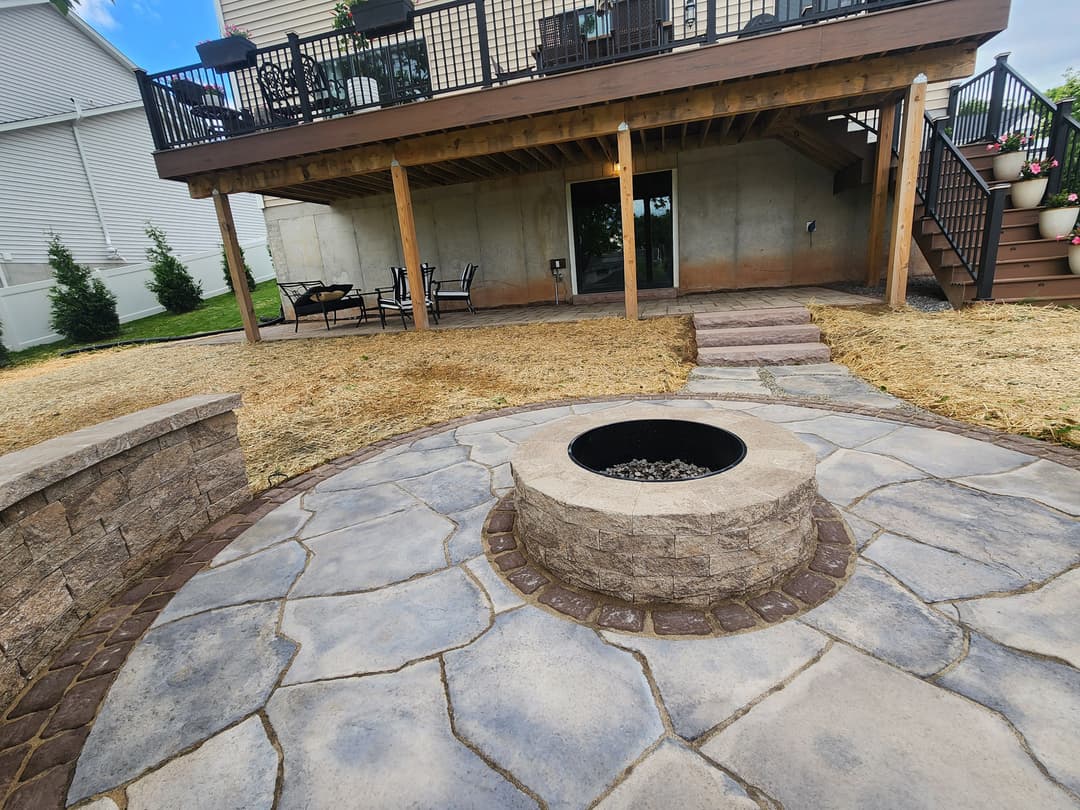 Backyard fire pit area with stone patio, wooden deck, and seating under a house.