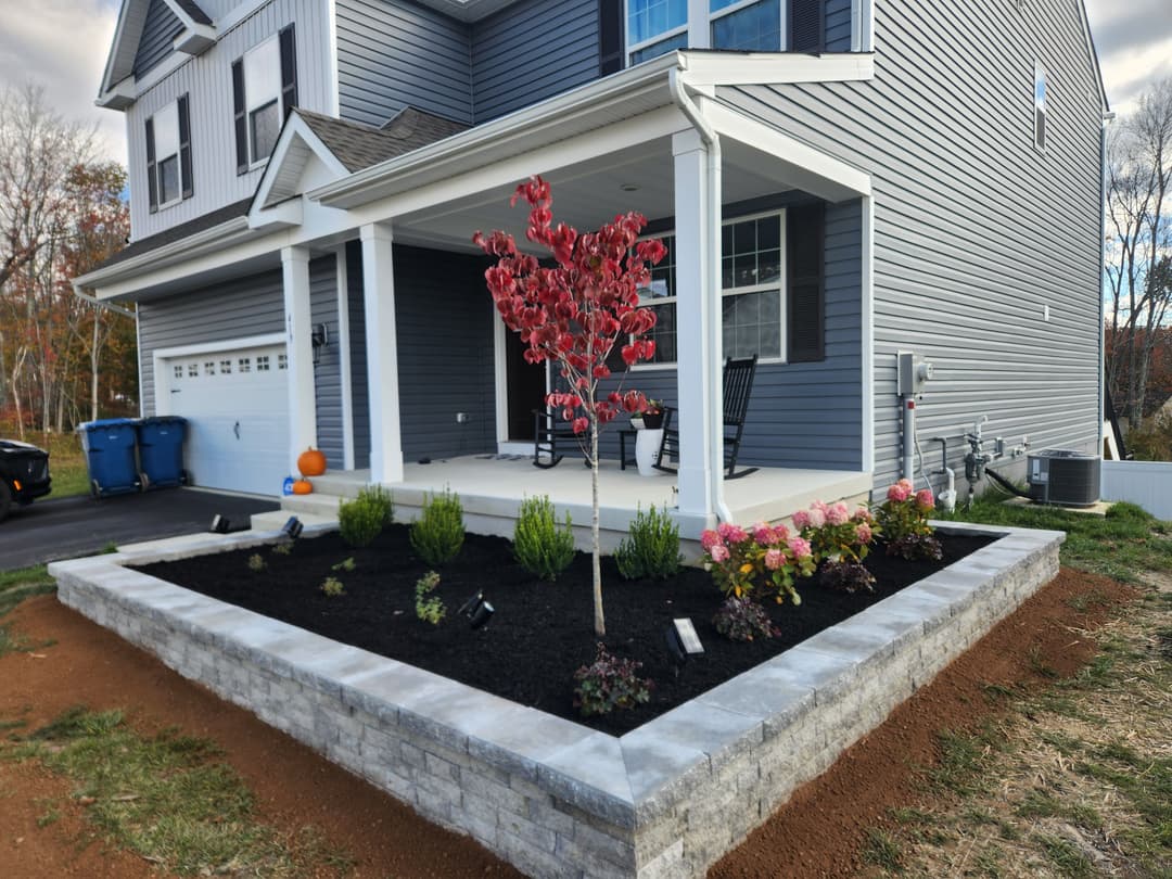 Lush garden with blooming flowers and a stone path beside a gray house on a sunny day.