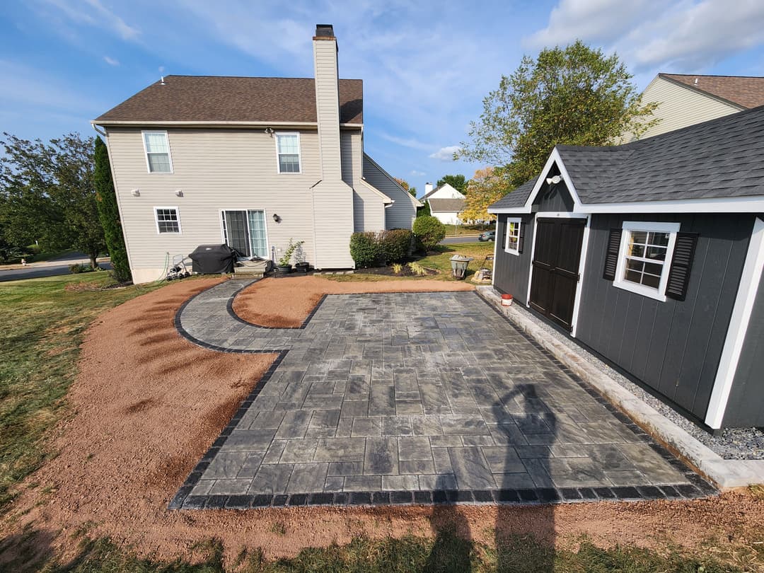 Paved patio and landscaping beside a house and shed in a residential backyard.