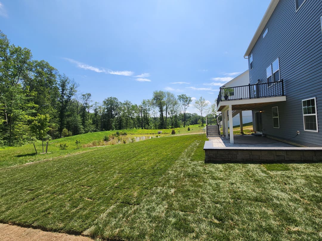 Lush green yard with freshly laid sod, scenic pond, and blue sky behind a modern home.