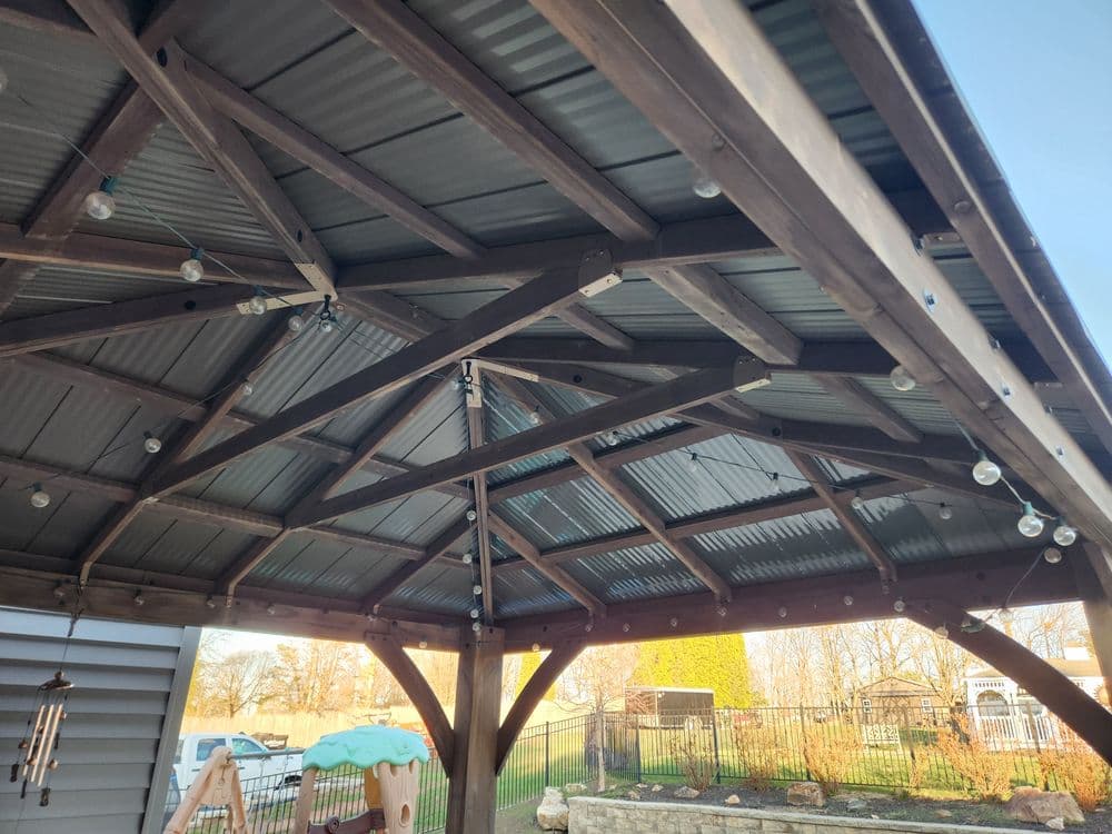 Wooden pergola with string lights and corrugated metal roof in a backyard setting.