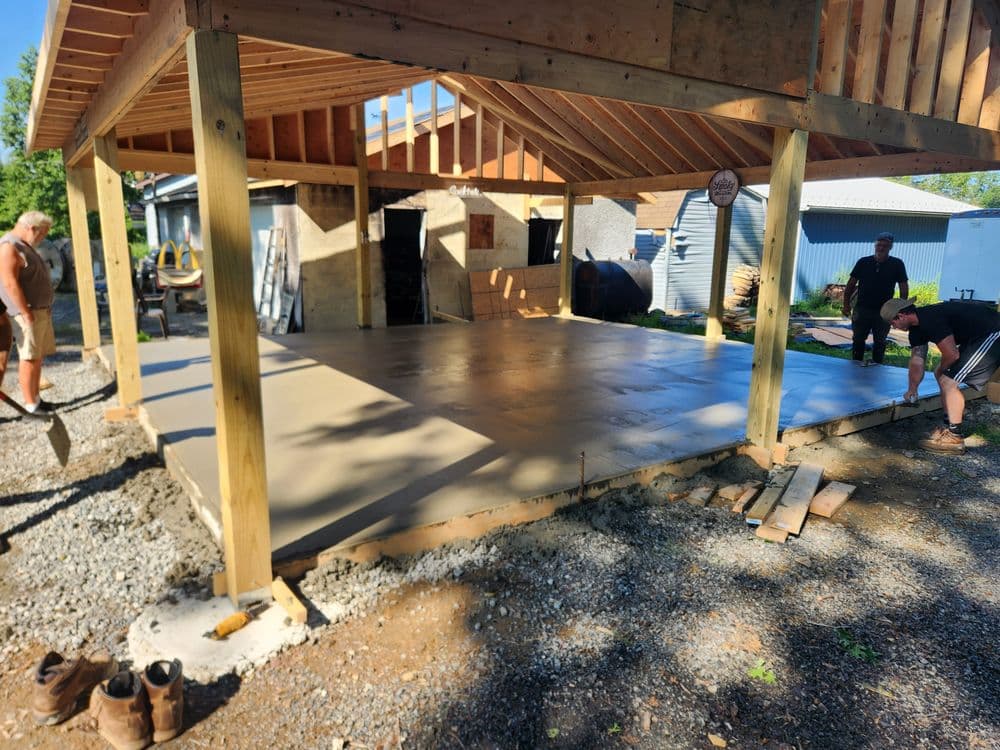 Construction workers leveling a concrete slab for a new outdoor structure under a wooden frame.
