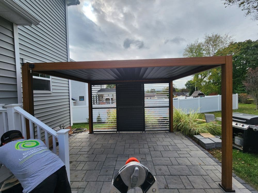 Modern outdoor patio pergola with vertical slats, gray pavers, and nearby landscaping.