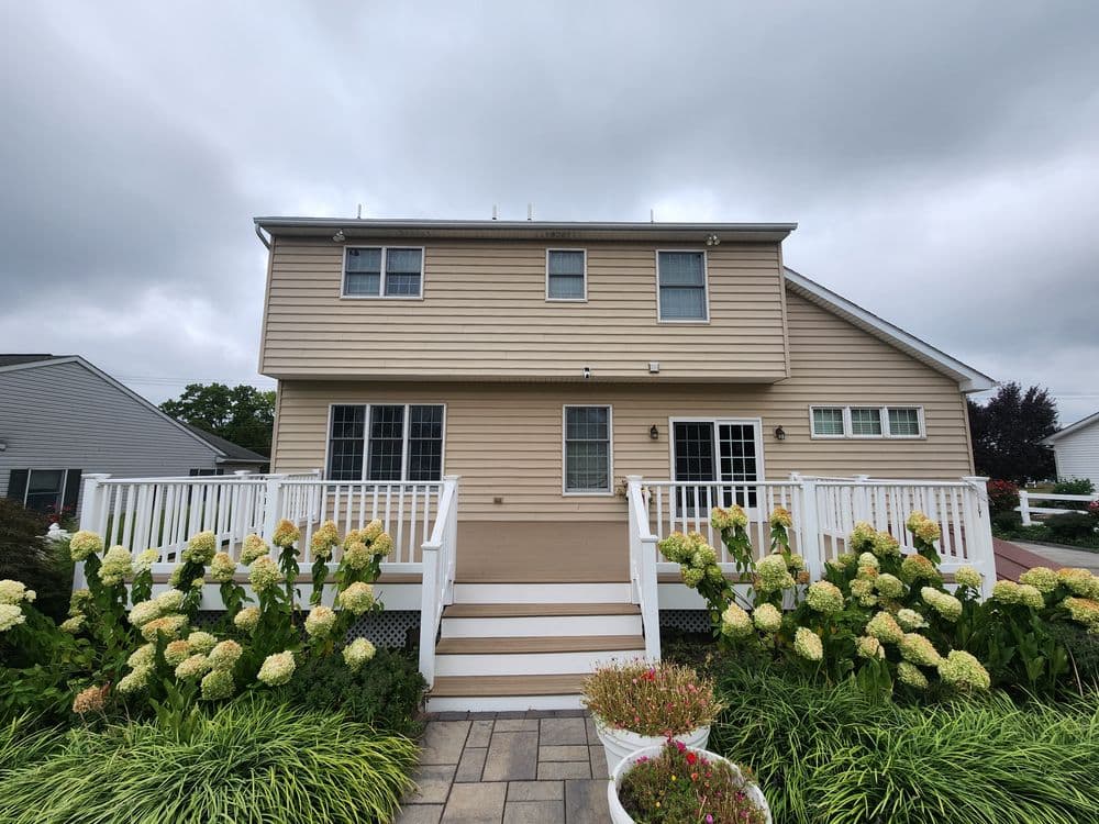 Two-story light brown house with white railings, surrounded by green hydrangeas and cloudy sky.