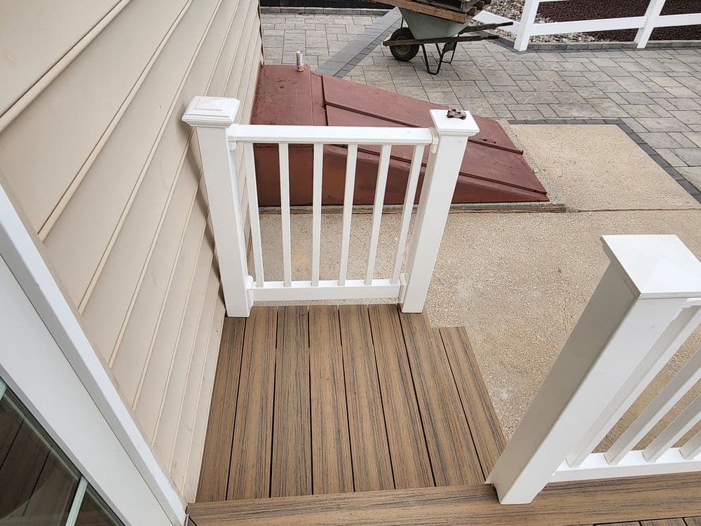 Wooden deck with white railing leading to a paved area and wheelbarrow in the background.