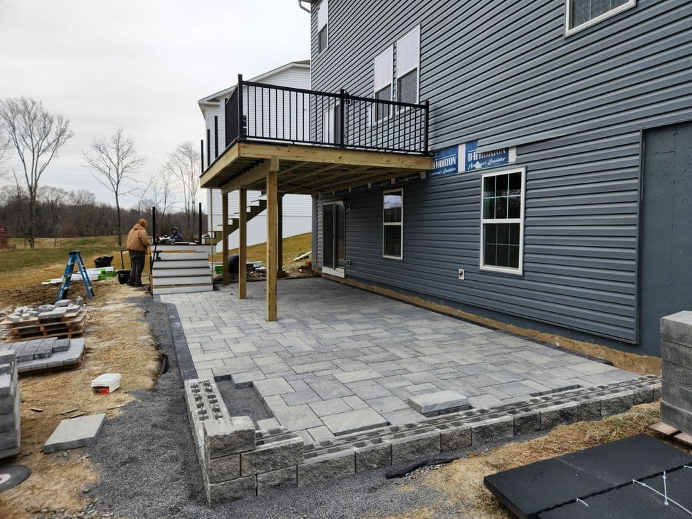 Paved patio installation under a deck with construction tools and materials in the background.