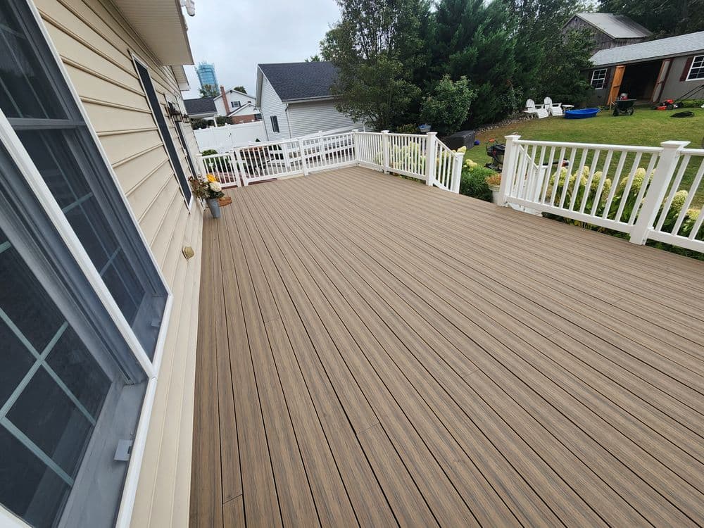 Spacious outdoor deck with wooden planks and white railing, surrounded by greenery and homes.