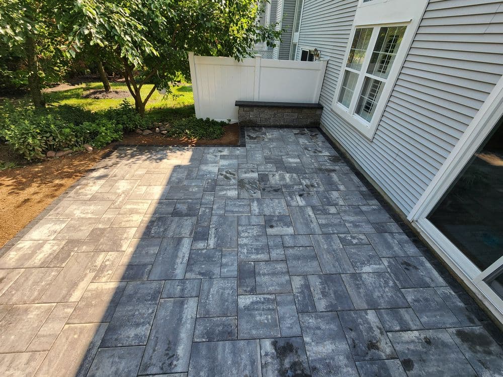 Outdoor patio with gray pavers, surrounded by greenery and a white fence.
