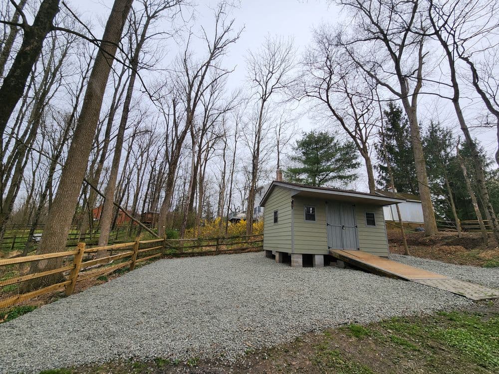 Small green shed on a gravel area surrounded by bare trees and a wooden fence in a wooded setting.