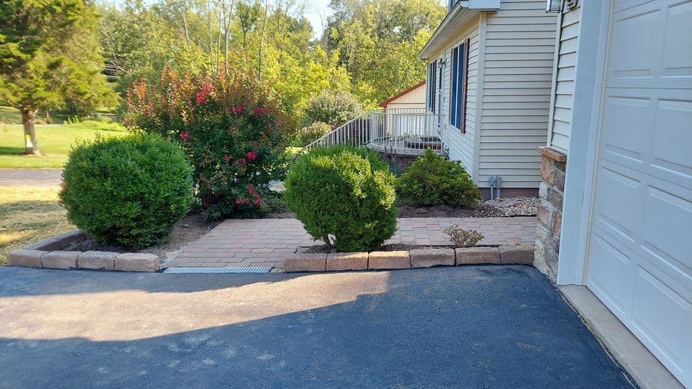 Lush garden pathway with shrubbery and blooming flowers beside a home's garage entrance.