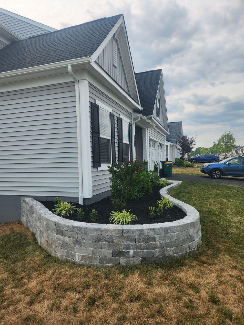 Landscaped home corner with stone edging, flowers, and gray siding under cloudy sky.
