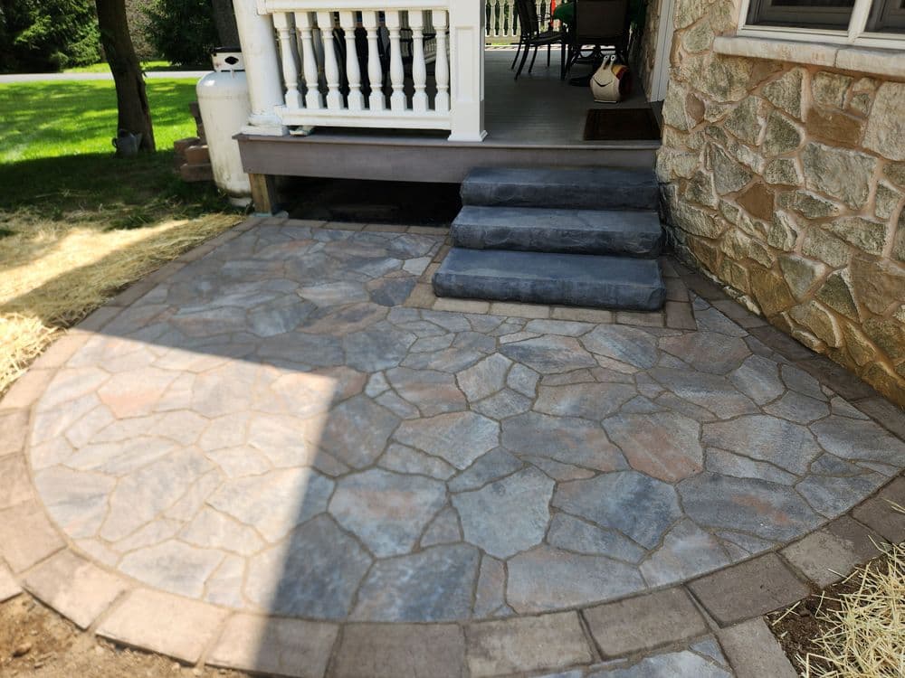 Stone patio with steps leading to a house, surrounded by grass and landscaping.