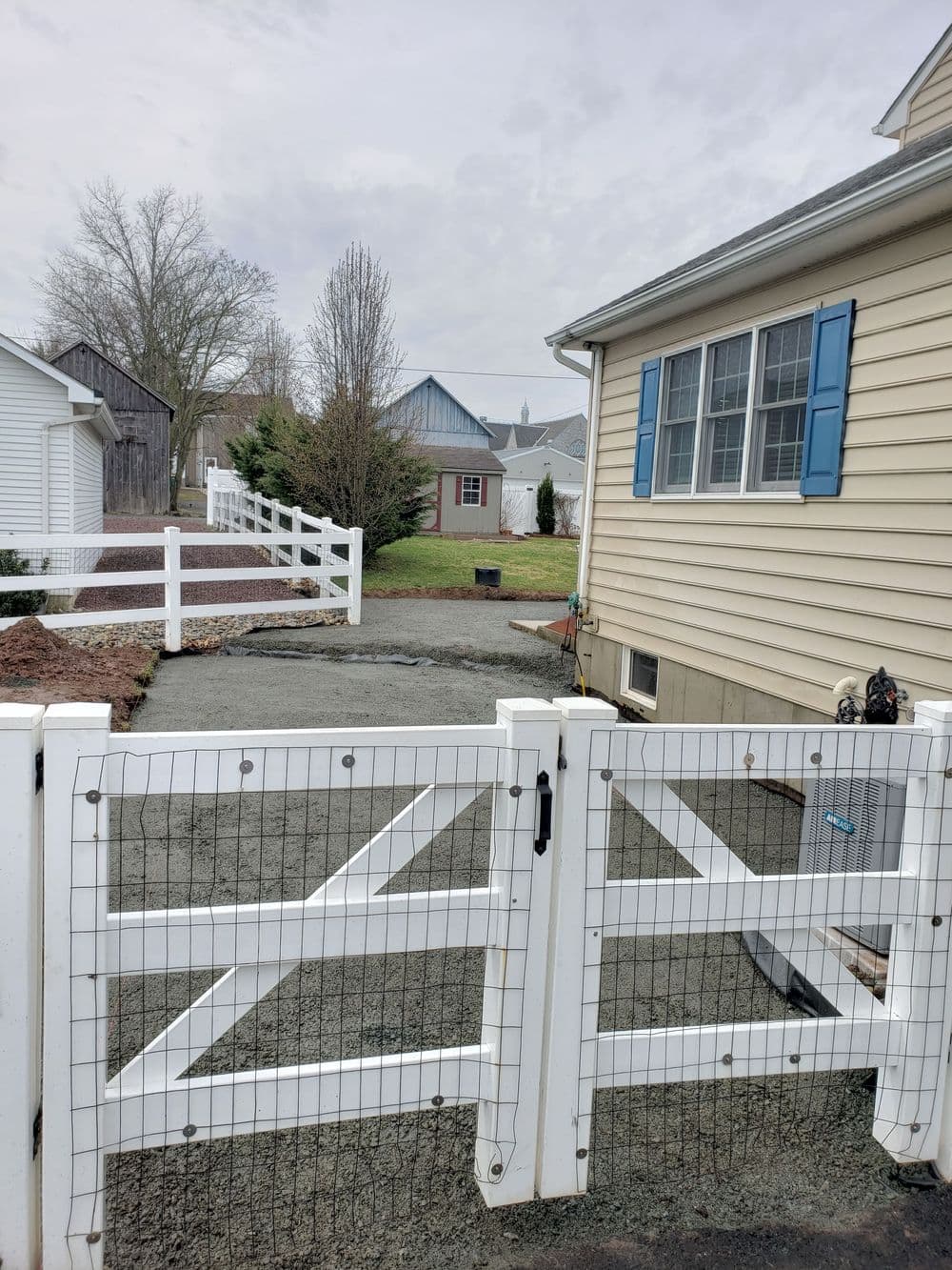 White fenced yard with gravel path, siding house, and a glimpse of greenery beyond the gate.