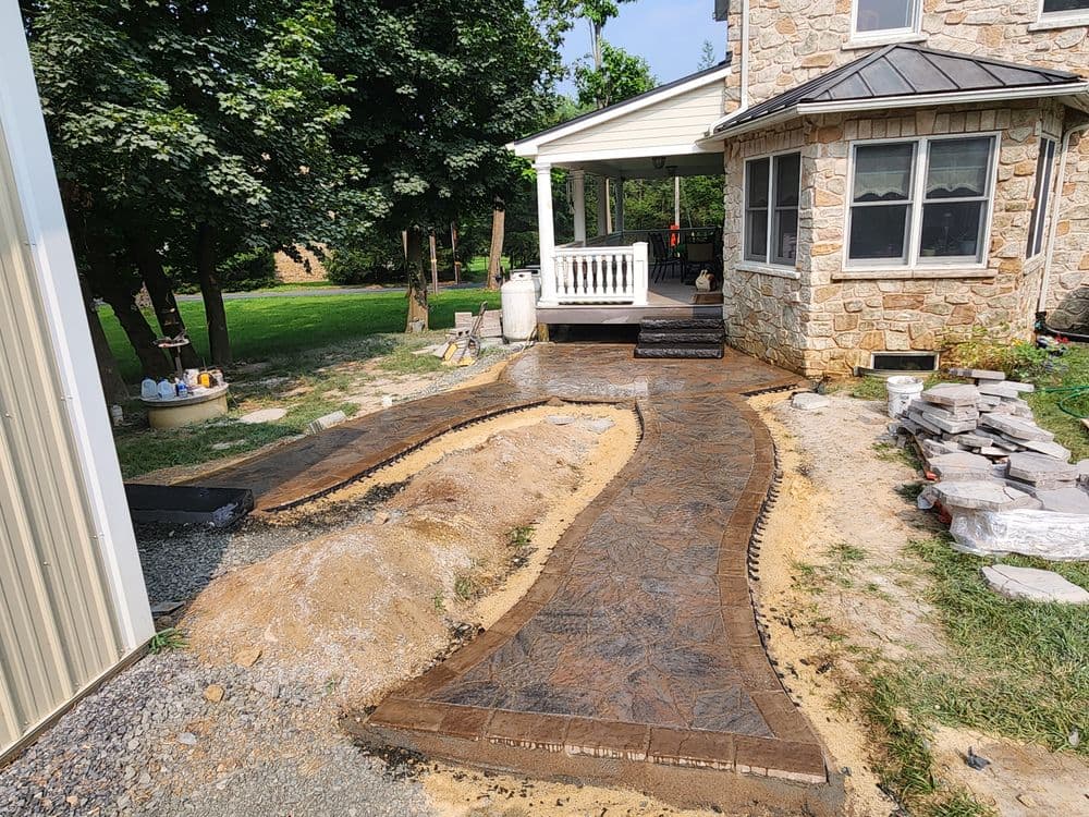 Decorative stamped concrete walkway leading to a house with stone exterior and lush greenery.