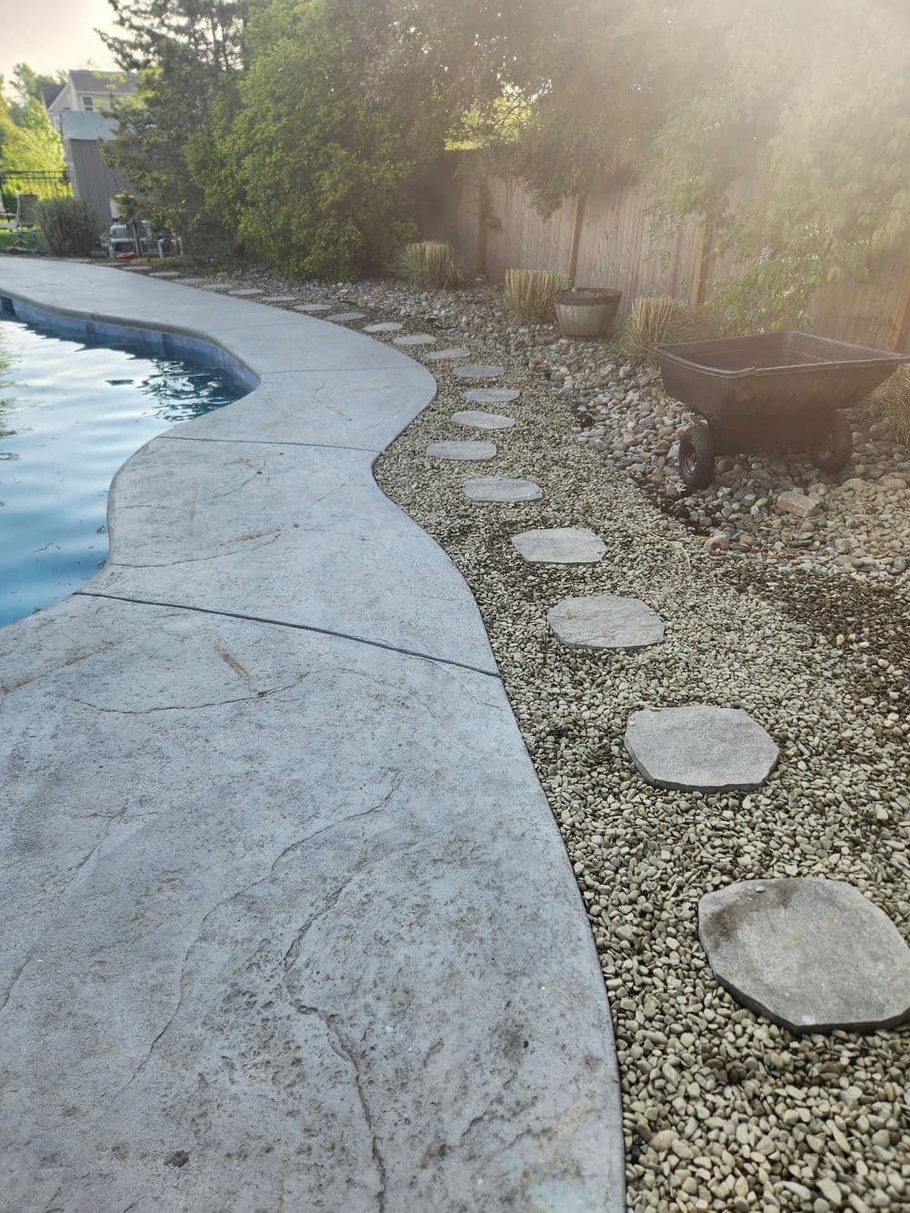 Stepping stones along a poolside path with gravel and a wheelbarrow nearby.