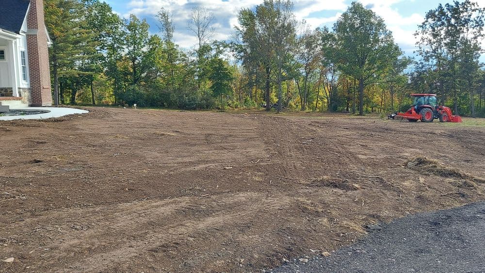 Cleared land near a house with a tractor and scattered trees under a blue sky.