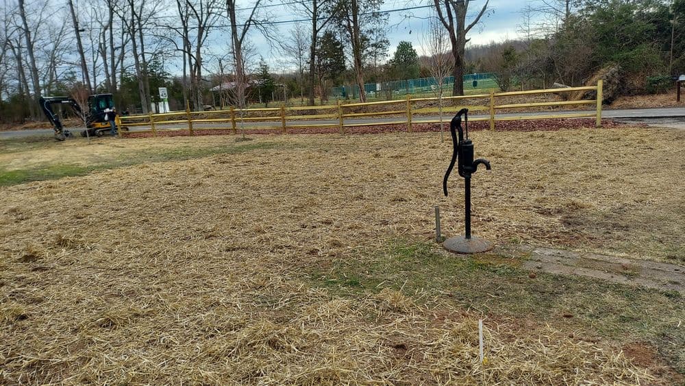Old-fashioned water pump on a straw-covered ground, with a wooden fence and machinery in the background.