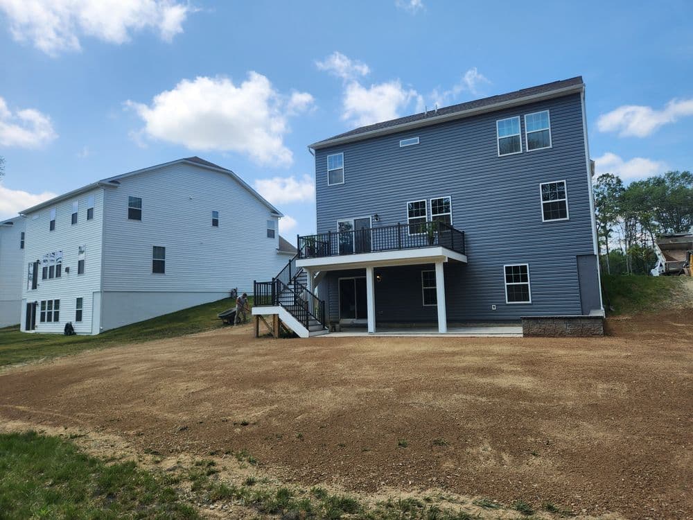 Newly built gray house with a large deck and gravel yard, surrounded by neighboring homes.