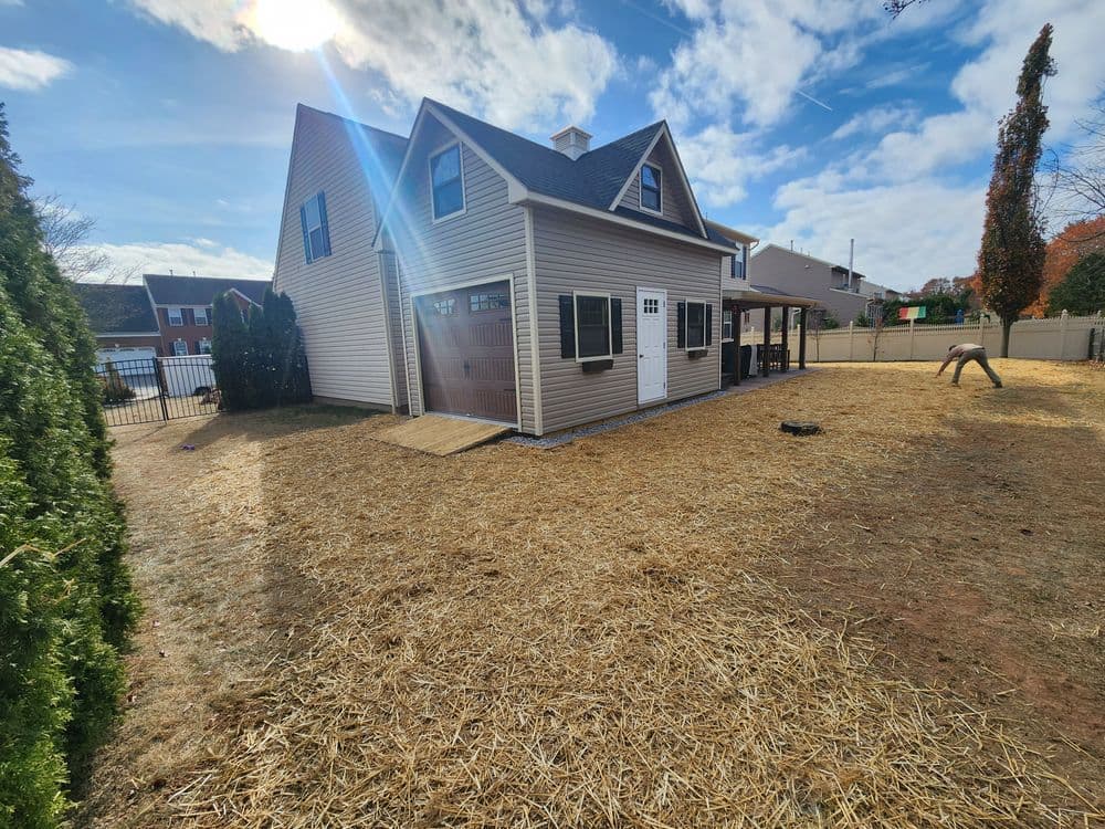 Backyard view of a two-story house with a wooden deck on a sunny day. Dry grass and fence visible.