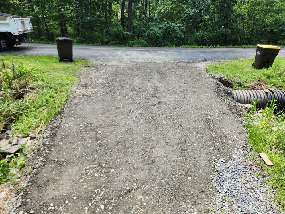 Gravel driveway leading to a road, surrounded by greenery and trash bins nearby.