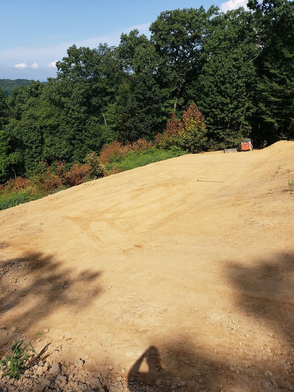 Cleared dirt site prepared for construction in a wooded area with trees and hills.