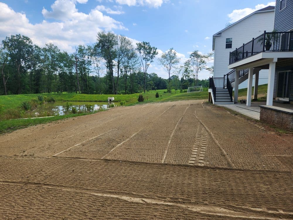 Cleared sandy area beside a home, with trees and a pond in the background under a blue sky.