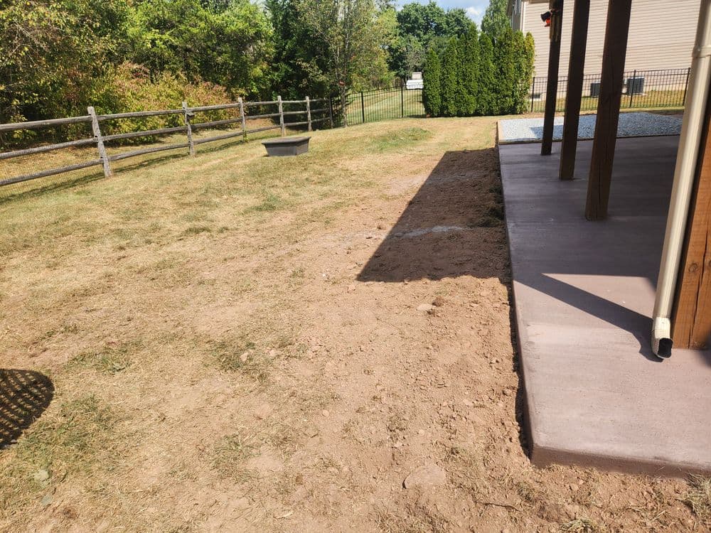Dry backyard area with dirt, grass patches, and a concrete patio near a wooden fence.