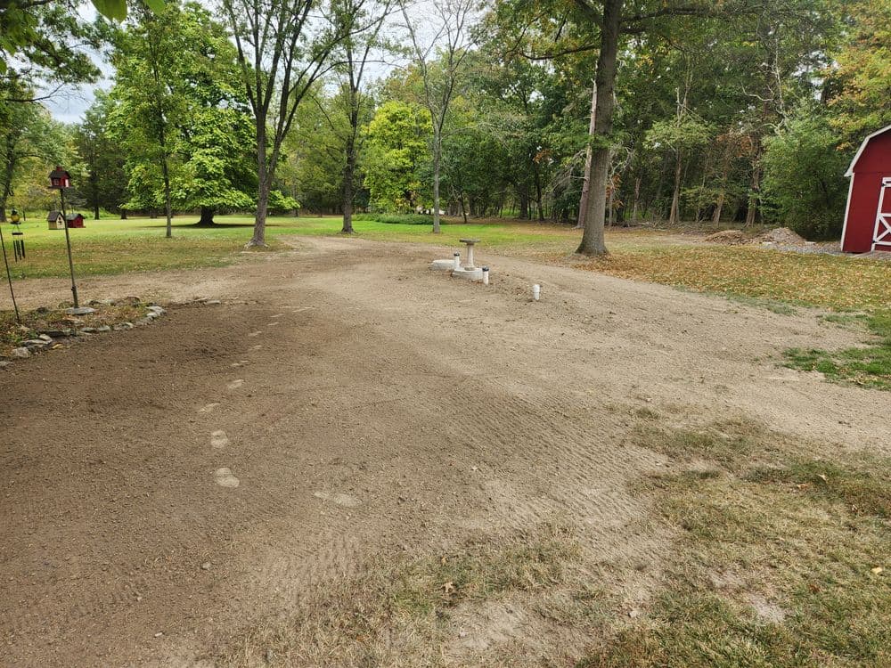 Cleared sandy area in a park with trees, bird feeder, and red barn in the background.