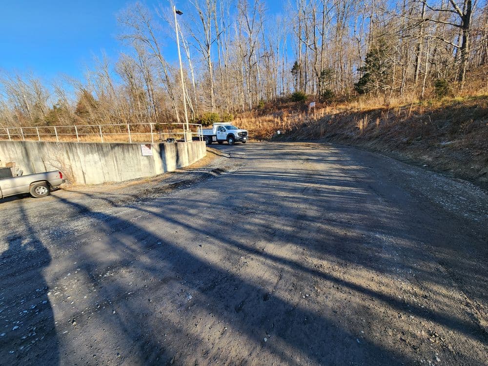 Gravel road leading to a parking area with vehicles and wooded surroundings.